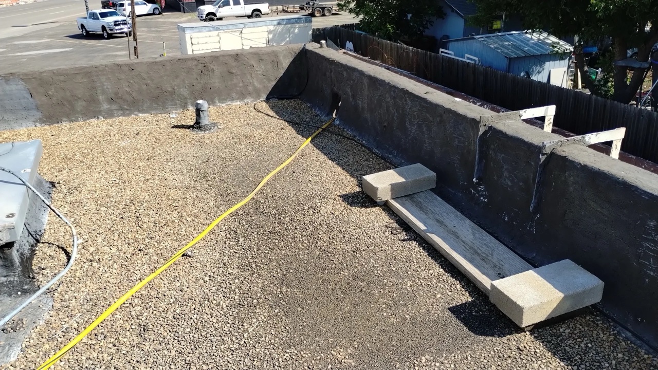 A flat commercial roof under construction showing built-up roofing with gravel surfacing partially installed, with a yellow