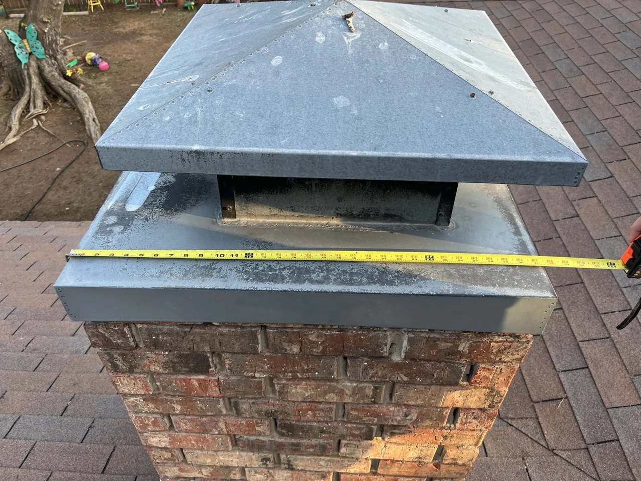 A metal chimney cap with protective flashing installed on top of a brick chimney, with a measuring tape extended across it